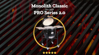 Monolith Classic PRO Series 2.0 Keramikgrill mit geöffnetem Deckel auf einem Gestell, umgeben von einem goldenen Kreis mit fünf Sternen Bewertung, vor glühenden Holzkohlen.