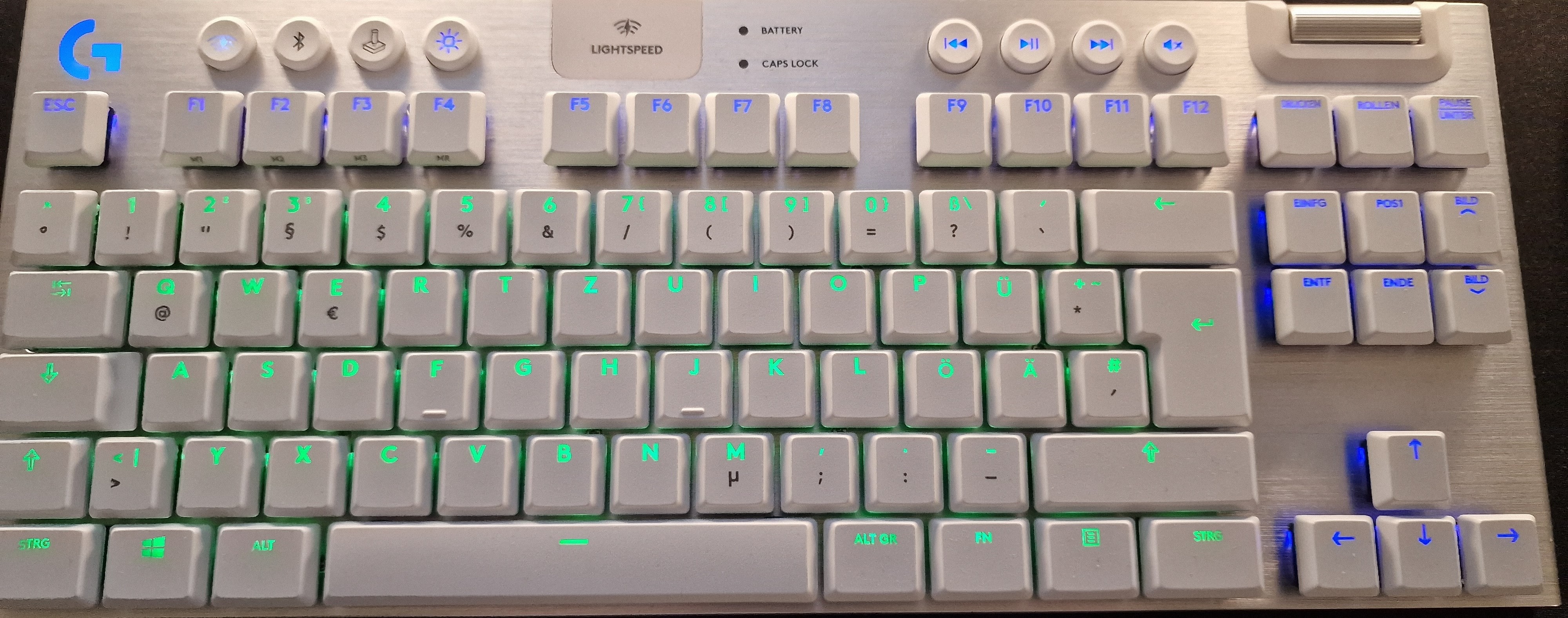 Das Bild zeigt eine Logitech G915 LIGHTSPEED TKL kabellose, Gaming Tastatur. Sie besitzt weiße Tastenkappen, die mit einer grünen Hintergrundbeleuchtung versehen sind, was einen ansprechenden Kontrast zum metallischen Finish des Tastaturkörpers bildet. Die Tastatur verfügt über dedizierte Mediensteuerungstasten im oberen rechten Bereich sowie ein metallenes Lautstärkerad. Die Tastenanordnung ist im TKL-Format (Tenkeyless), also ohne Nummernblock, was für mehr Platz auf dem Schreibtisch sorgt und bei Gamern beliebt ist. Die Beschriftungen der Tasten sind auf Deutsch, inklusive Umlauten und typischen Layout-Merkmalen wie "STRG" für Steuerungstaste.