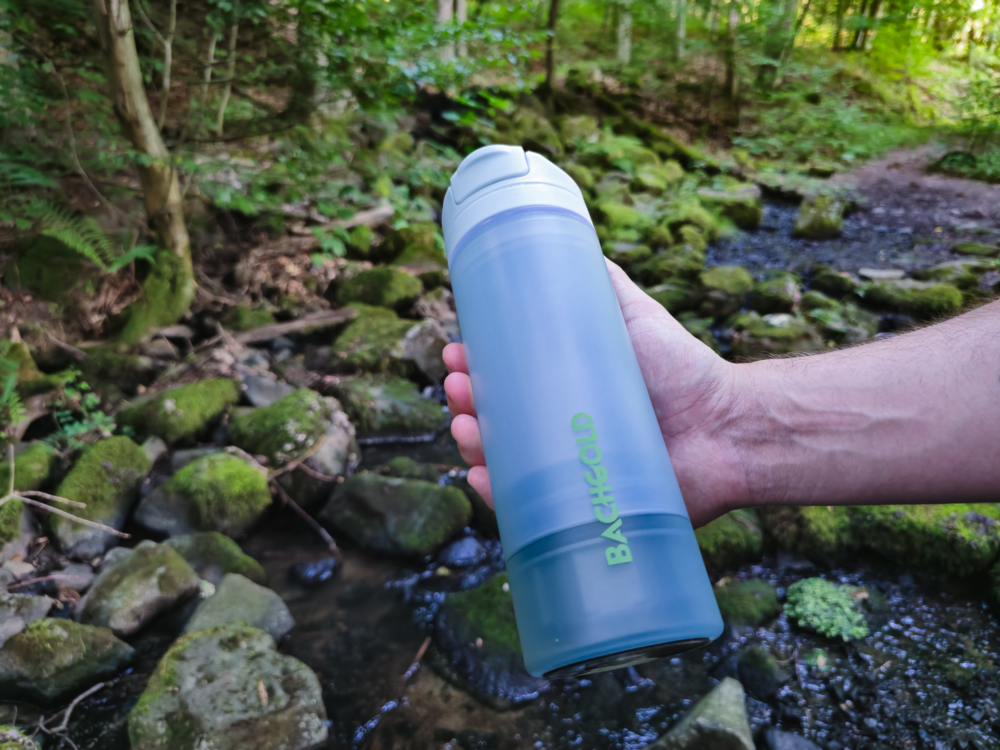 Person hält den Bachgold Outdoor Wasserfilter in der Hand, umgeben von einem Waldgebiet