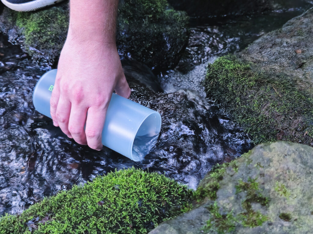 Person entnimmt Wasser aus einem Bach mit dem Äußerern Behälter