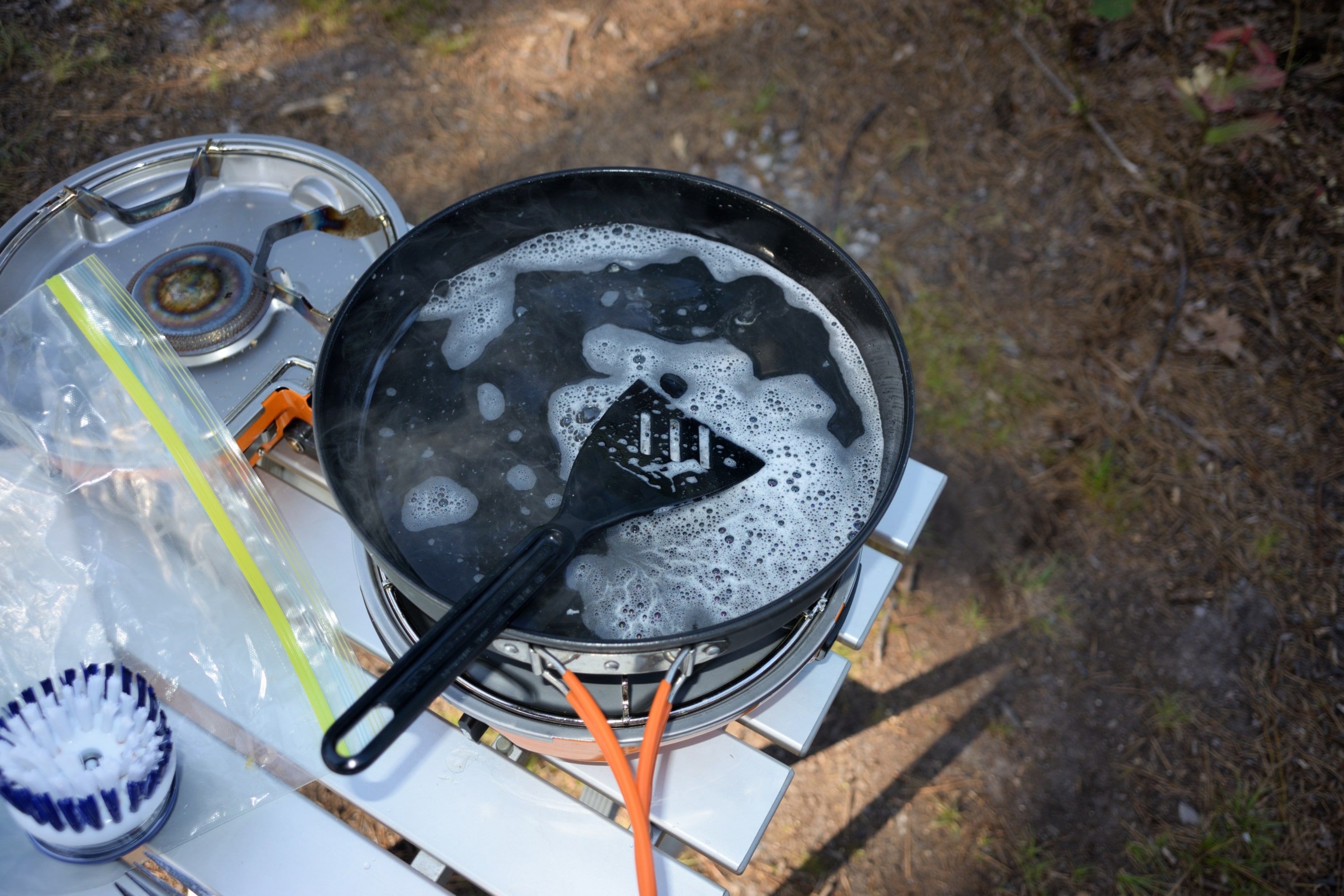 Runde Camping-Grillplatte beim Abwasch auf Einflamm-Gaskocher – Outdoor-Einsatz, Seifenwasser & Pfannenwender zeigen unkomplizierte Reinigung unterwegs.