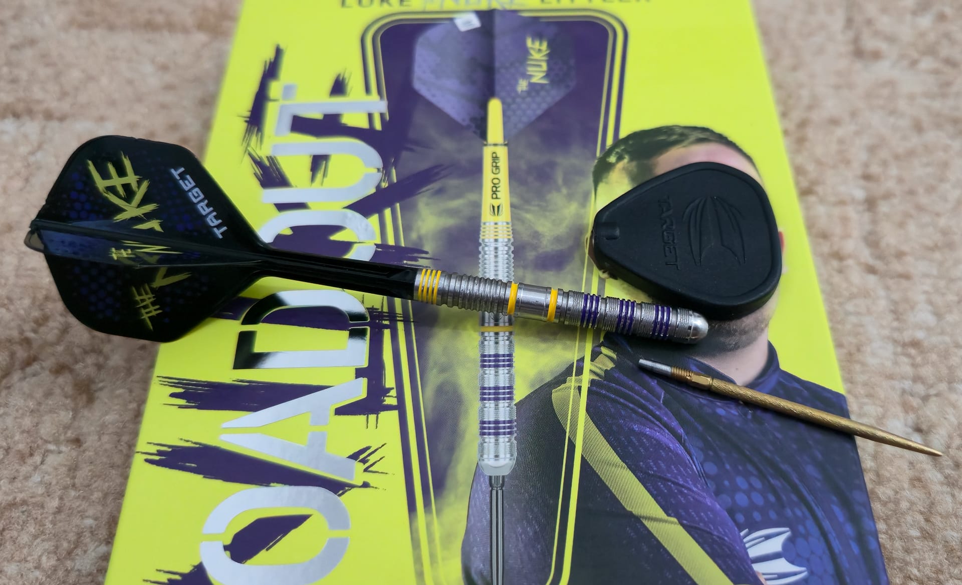 Luke Littler Loadout Dart liegt auf gelber Verpackung zusammen mit Swiss-Point-Werkzeug und Ersatzspitze; komplettes Set sichtbar.	