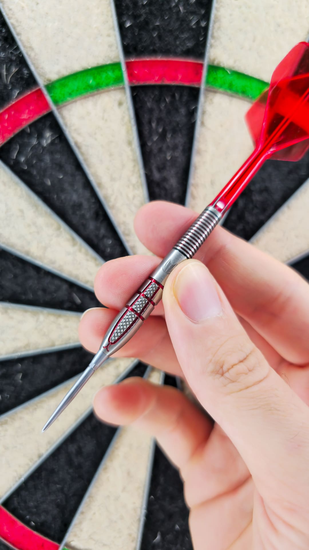 Nahaufnahme einer Hand, die einen roten Target Darts Nathan Aspinall Gen 2 Dart (95 % Wolfram, 26 g) an seinem griffigen Lauf hält, im Hintergrund unscharf eine traditionelle Sisal-Dartscheibe.
