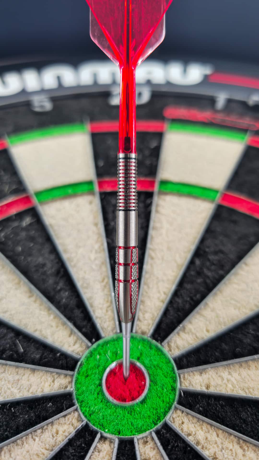 Frontale Aufnahme eines Nathan Aspinall Gen 2 Darts mit rotem Flight, der genau im roten Zentrum (Bull) der Dartboard-Scheibe steckenbleibt.