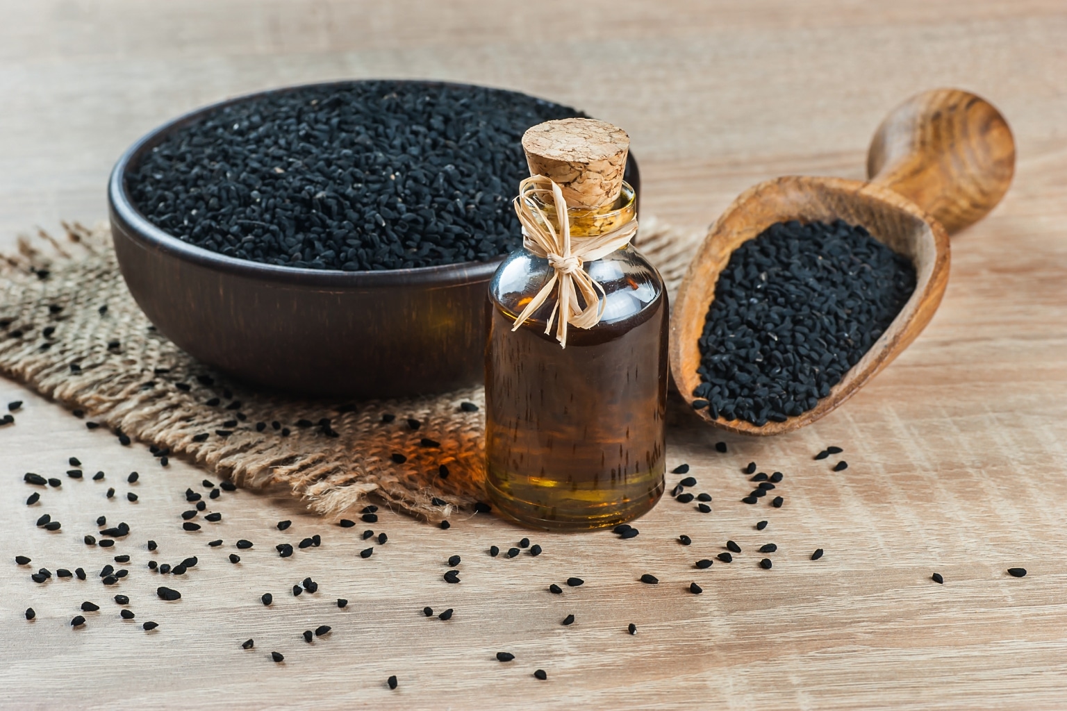 Schwarzkümmelöl für Babys – Fläschchen mit Schwarzkümmelöl vor einer Schale und einem Holzlöffel mit Schwarzkümmelsamen auf Holzuntergrund.