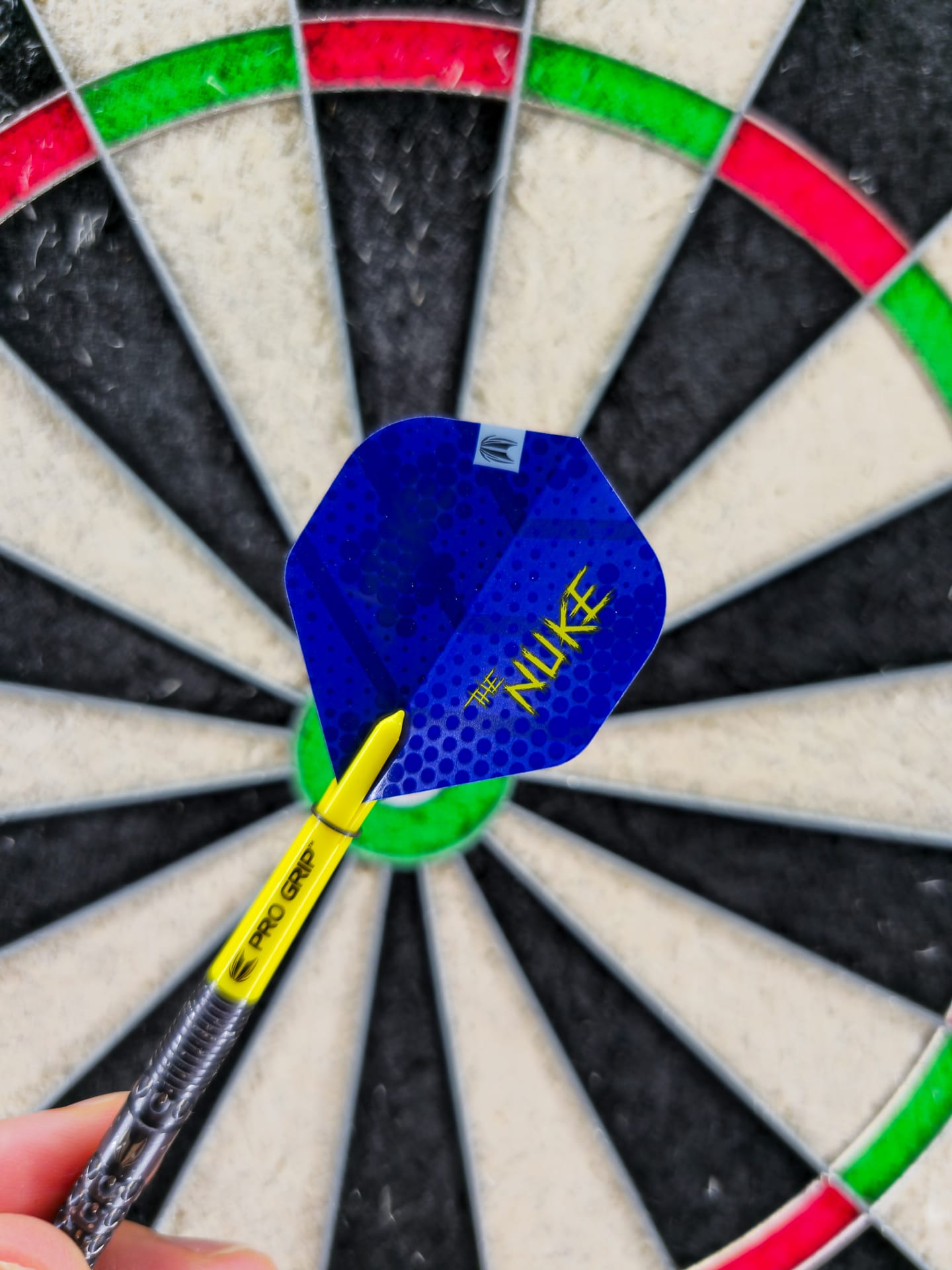 Hand hält Luke Littler Dart mit gelbem Pro-Grip-Shaft und blauem Nuke-Flight vor dem Bull