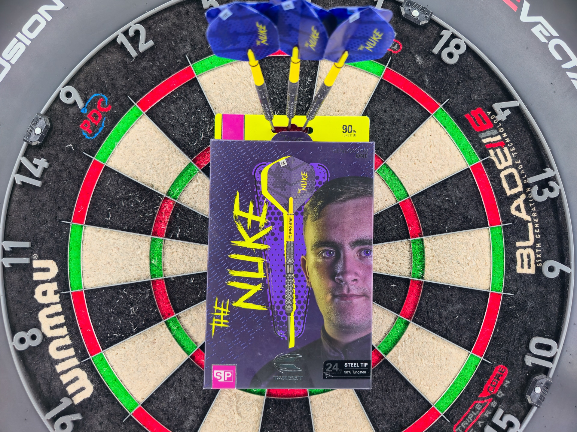 Luke Littler G1 SP „The Nuke“ Darts-Verpackung im Bulls-Eye einer Winmau Blade 6, blau-gelbe Flights gruppiert darüber