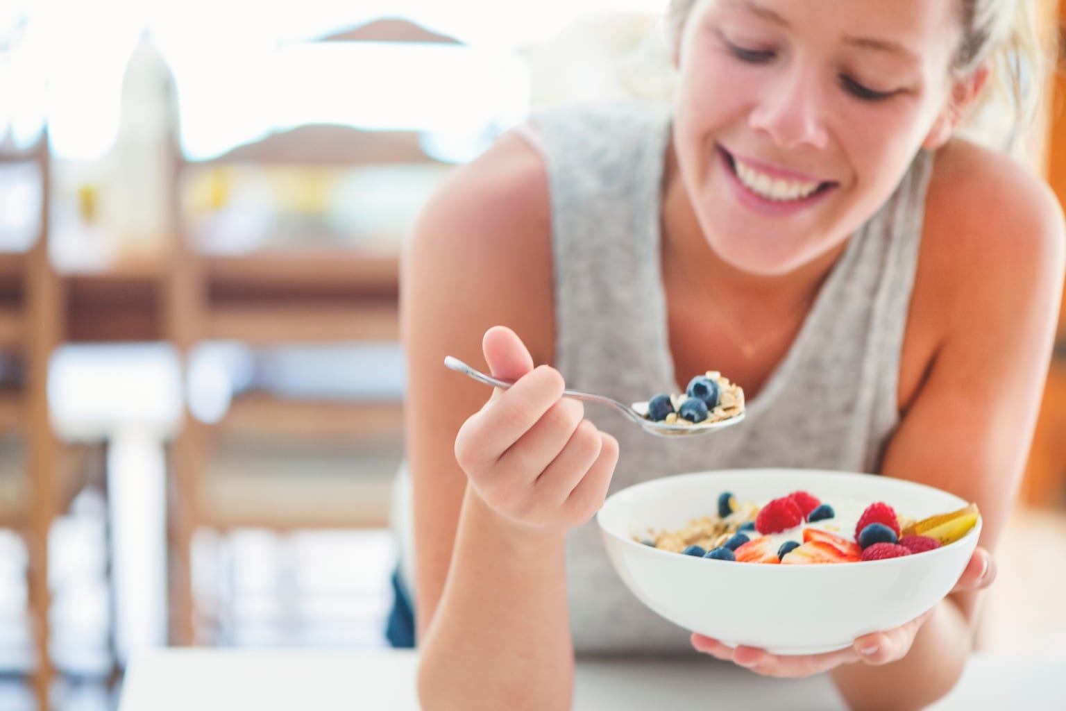 Eine lächelnde Frau isst eine Frühstücksschüssel mit Haferflocken, Beeren und Früchten, symbolisierend eine gesunde Akne Inversa Ernährung.